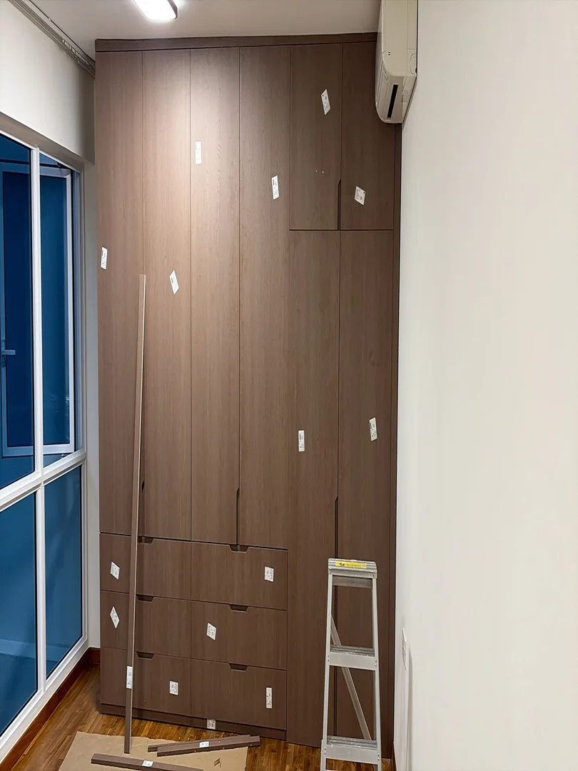 Tall brown built-in wardrobe being installed beside a bedroom window and air-conditioner in a Singapore HDB home